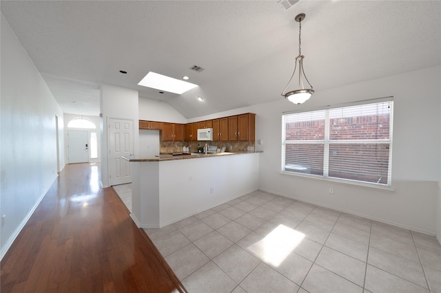 kitchen featuring vaulted ceiling with skylight, white microwave, a peninsula, brown cabinets, and decorative backsplash