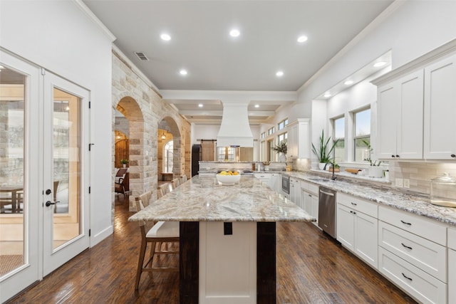 kitchen with custom range hood, dark wood-type flooring, light stone countertops, dishwasher, and a kitchen bar