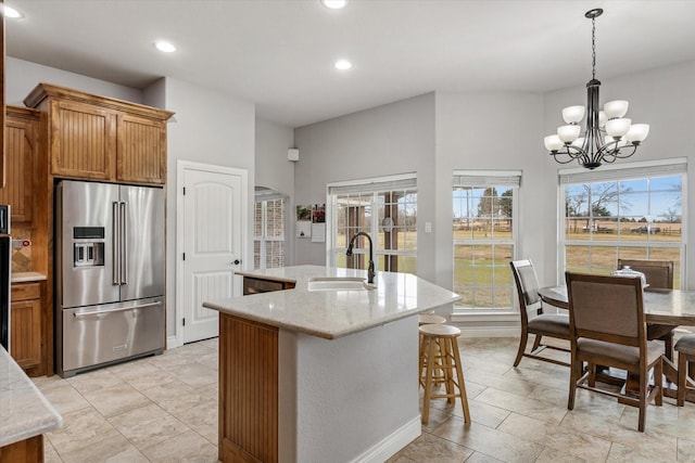 kitchen with high end refrigerator, a sink, brown cabinetry, an island with sink, and an inviting chandelier