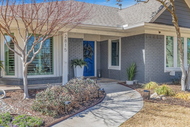 view of exterior entry with a shingled roof, brick siding, and a porch