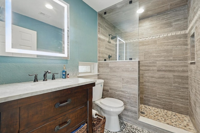 full bath featuring visible vents, toilet, a walk in shower, vanity, and a textured wall