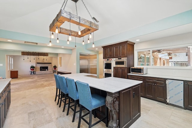 kitchen with stainless steel appliances, a fireplace with raised hearth, a kitchen bar, dark brown cabinetry, and a kitchen island with sink