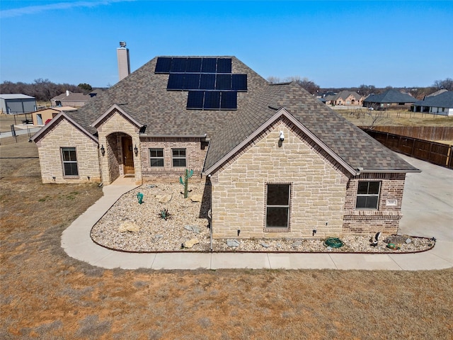 french country style house with solar panels, a shingled roof, brick siding, stone siding, and a chimney