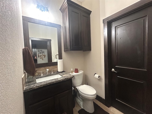 half bath featuring baseboards, a textured wall, vanity, and toilet