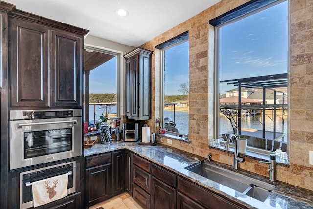 kitchen featuring dark brown cabinetry, recessed lighting, a water view, a sink, and dark stone counters