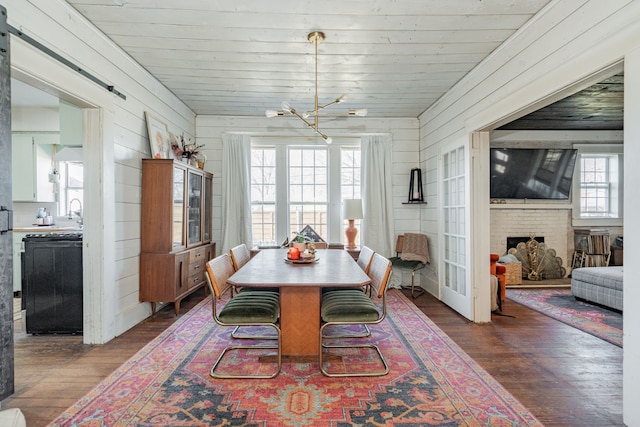 dining area with dark wood-type flooring, a brick fireplace, wooden walls, and an inviting chandelier