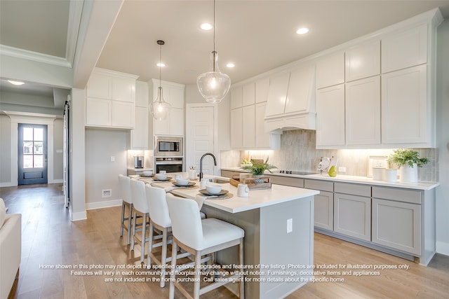 kitchen with light wood finished floors, decorative backsplash, a kitchen island with sink, stainless steel appliances, and premium range hood