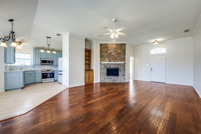 unfurnished living room featuring crown molding, a stone fireplace, light wood-type flooring, a sink, and ceiling fan with notable chandelier