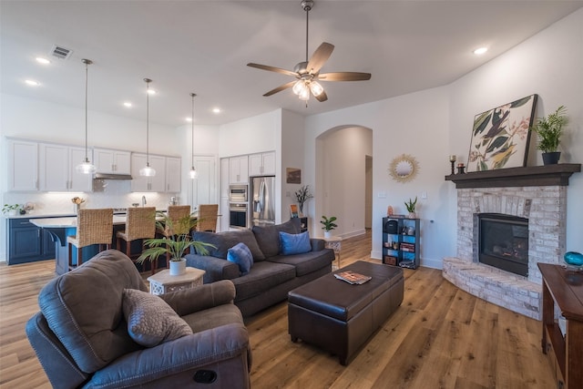 living area with arched walkways, visible vents, light wood-style flooring, ceiling fan, and a stone fireplace