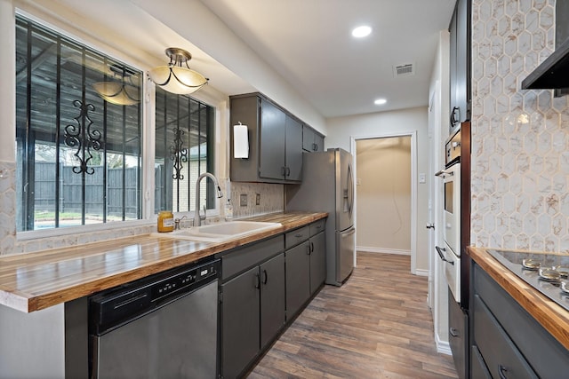 kitchen featuring decorative backsplash, butcher block countertops, stainless steel appliances, wall chimney range hood, and a sink