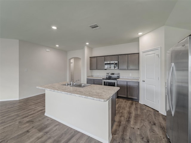 kitchen featuring arched walkways, dark wood-type flooring, a sink, appliances with stainless steel finishes, and an island with sink