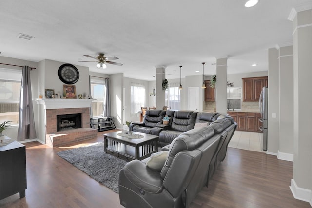 living area featuring light wood-style flooring, visible vents, baseboards, a brick fireplace, and ornate columns