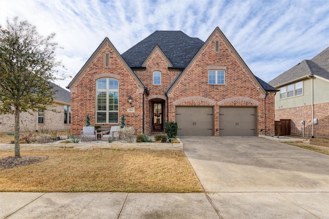 english style home with a garage, concrete driveway, brick siding, and a shingled roof