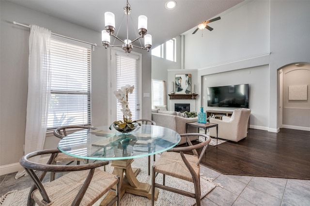 dining space featuring light tile patterned floors, a towering ceiling, a glass covered fireplace, baseboards, and ceiling fan with notable chandelier