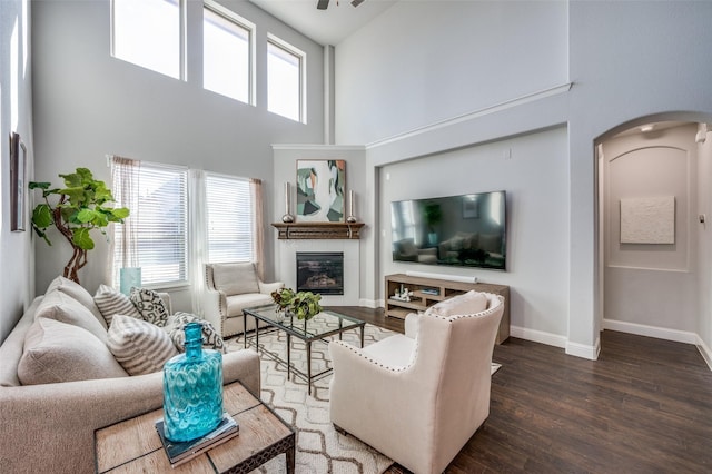 living room featuring arched walkways, a high ceiling, a glass covered fireplace, wood finished floors, and baseboards