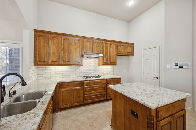 kitchen featuring under cabinet range hood, light stone counters, brown cabinetry, and a sink