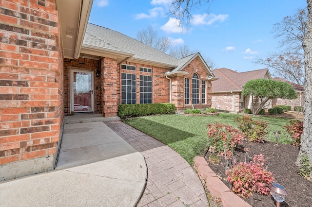 view of exterior entry with a lawn, brick siding, and a shingled roof