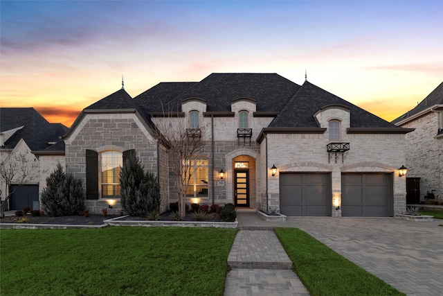 french country home with decorative driveway, roof with shingles, a lawn, an attached garage, and stone siding