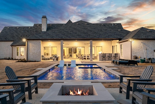 back of house at dusk featuring roof with shingles, a chimney, a patio area, an outdoor pool, and a fire pit