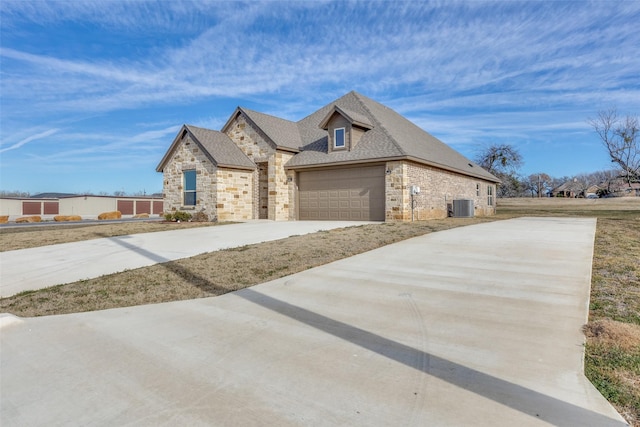 view of front of house featuring roof with shingles, an attached garage, central AC, stone siding, and driveway