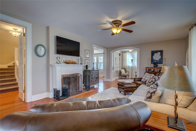 living room featuring arched walkways, ceiling fan, wood finished floors, stairs, and a brick fireplace