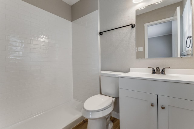 bathroom with toilet, a textured wall, a tile shower, and vanity