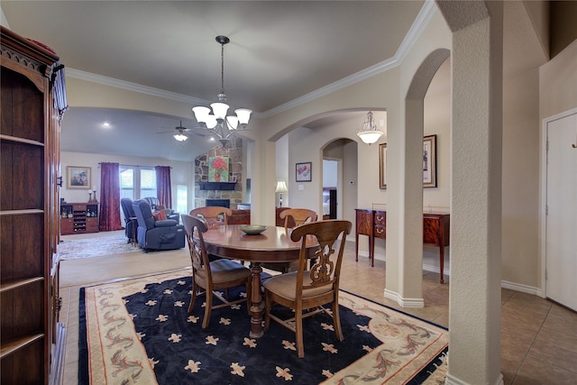 tiled dining area featuring arched walkways, crown molding, a stone fireplace, baseboards, and ceiling fan with notable chandelier