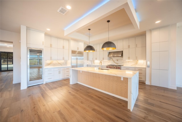 kitchen featuring light wood-style flooring, a tray ceiling, stainless steel built in refrigerator, a sink, and exhaust hood