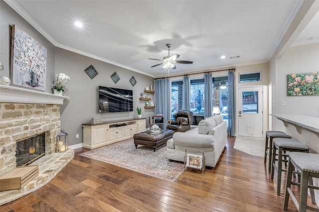 living room featuring a stone fireplace, dark wood-type flooring, a ceiling fan, baseboards, and ornamental molding