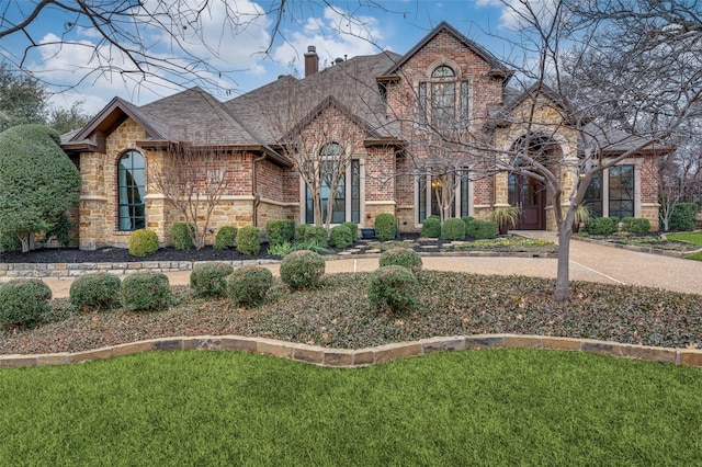 french country home with brick siding, stone siding, a chimney, roof with shingles, and a front yard