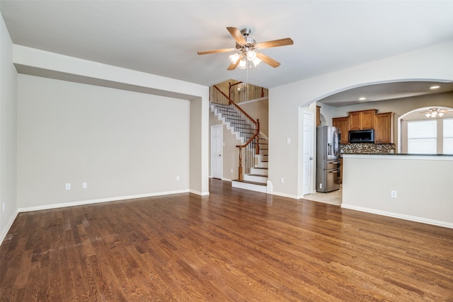 unfurnished living room with ceiling fan, arched walkways, wood finished floors, baseboards, and stairway