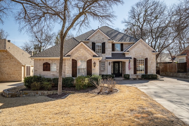 french country inspired facade with stone siding, brick siding, a shingled roof, and fence