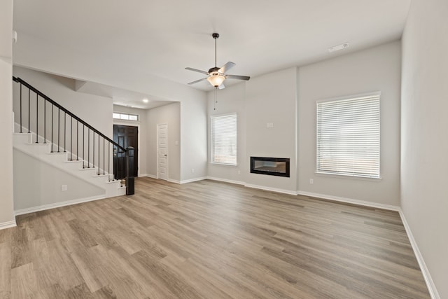 unfurnished living room featuring light wood-style flooring, stairway, a glass covered fireplace, ceiling fan, and baseboards