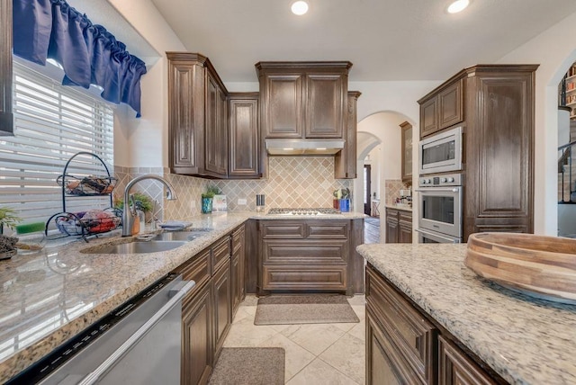 kitchen with light stone counters, under cabinet range hood, a sink, appliances with stainless steel finishes, and decorative backsplash