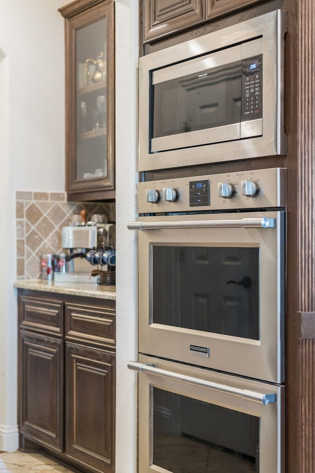 kitchen with light stone counters, stainless steel appliances, backsplash, glass insert cabinets, and dark brown cabinetry