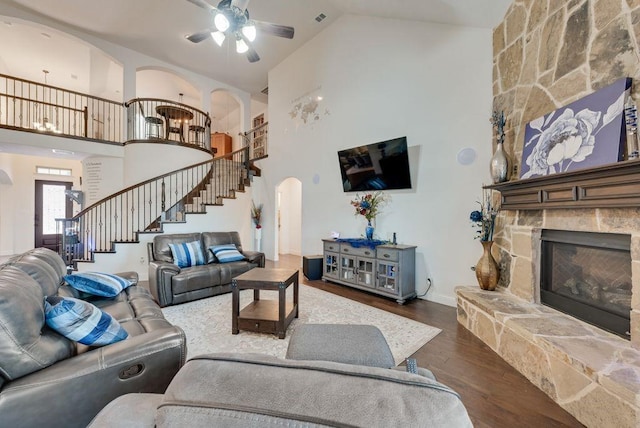 living room with arched walkways, wood finished floors, stairs, a stone fireplace, and high vaulted ceiling