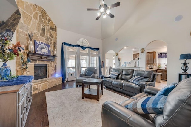 living area with arched walkways, a high ceiling, ceiling fan, a stone fireplace, and wood finished floors