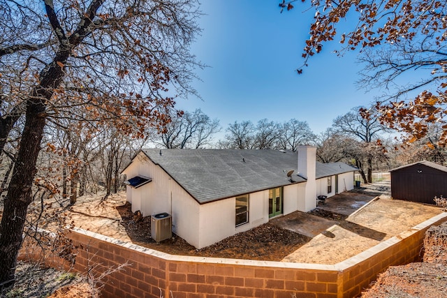 rear view of house featuring central air condition unit, a shingled roof, a chimney, and an outbuilding