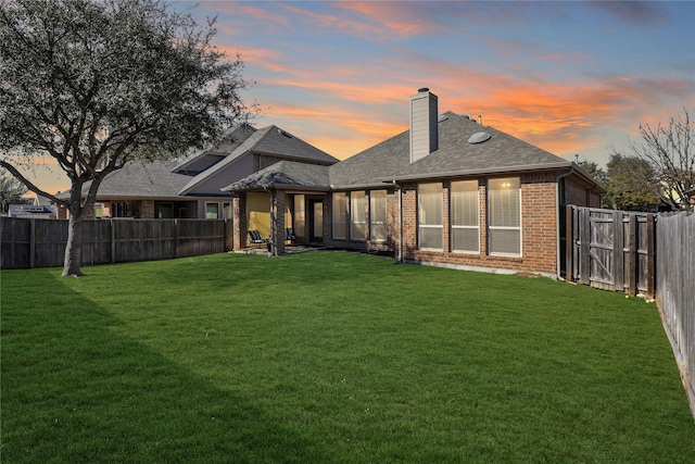 back of property at dusk featuring a fenced backyard, a chimney, a lawn, and brick siding