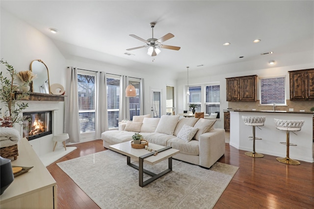 living area featuring dark wood-style floors, ceiling fan, visible vents, and a glass covered fireplace