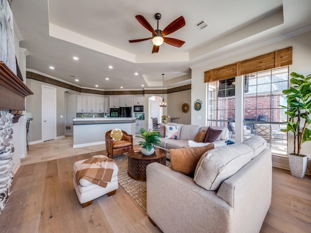 living area featuring ceiling fan with notable chandelier, visible vents, ornamental molding, a tray ceiling, and light wood finished floors