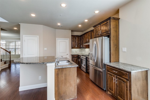 kitchen with a sink, an island with sink, stone counters, appliances with stainless steel finishes, and dark wood-type flooring