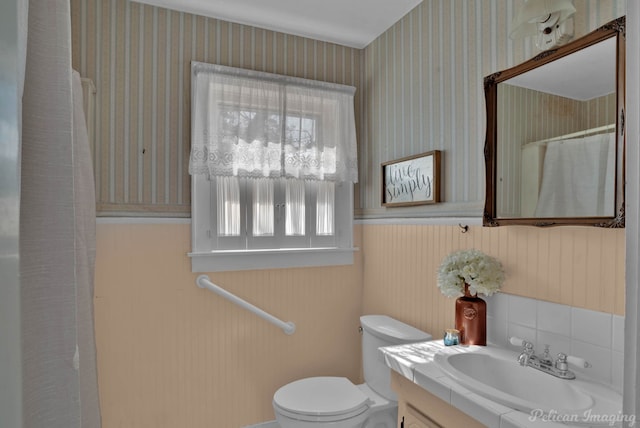 bathroom featuring wallpapered walls, a shower with shower curtain, toilet, a wainscoted wall, and vanity