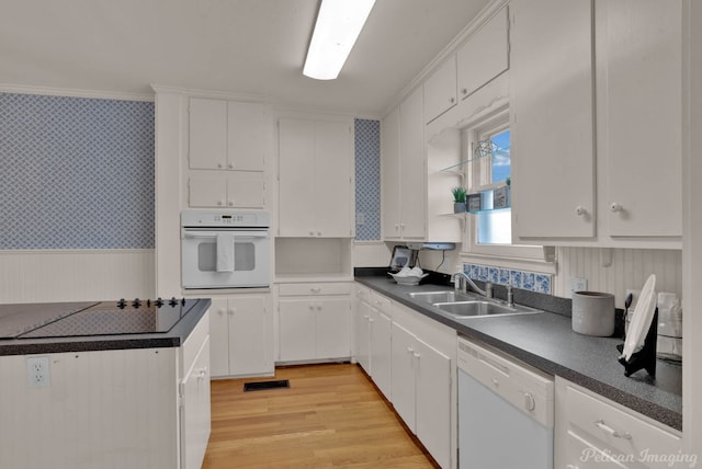 kitchen with white appliances, wallpapered walls, dark countertops, a wainscoted wall, and a sink