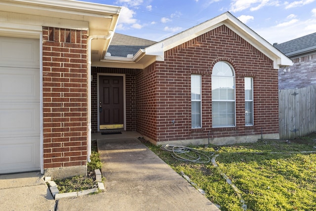 view of exterior entry featuring an attached garage, roof with shingles, fence, and brick siding