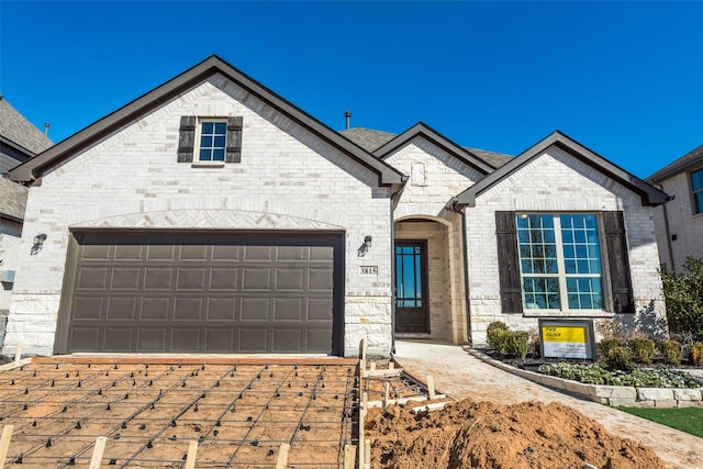 french country inspired facade featuring a shingled roof, brick siding, driveway, and an attached garage