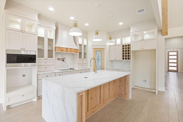 kitchen featuring black electric stovetop, a sink, visible vents, custom exhaust hood, and a center island with sink