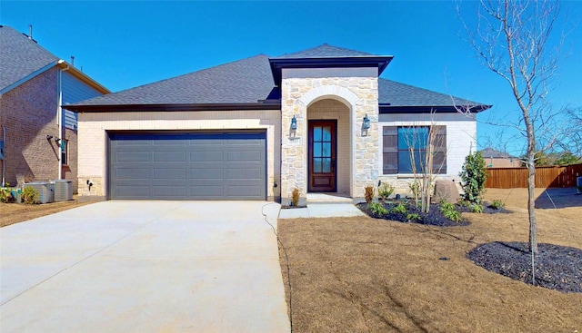 view of front of property with driveway, central AC unit, stone siding, roof with shingles, and an attached garage