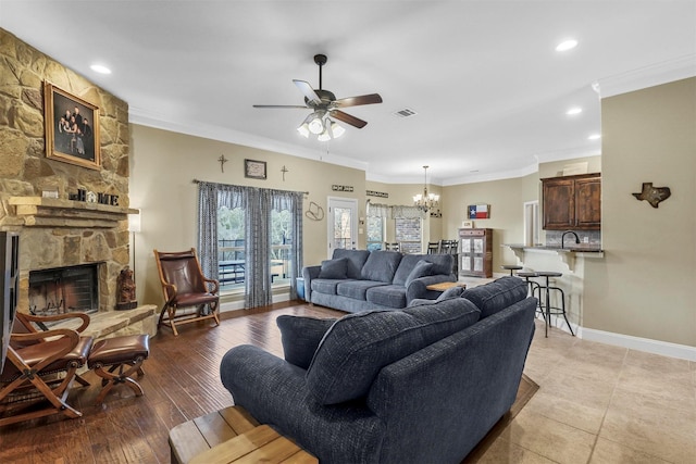 living room featuring visible vents, ornamental molding, a stone fireplace, baseboards, and ceiling fan with notable chandelier