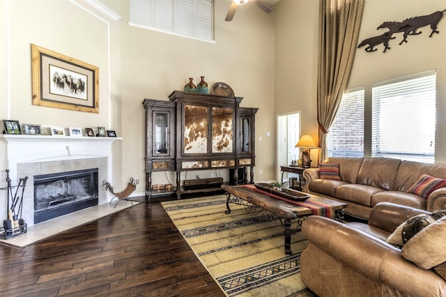 living room with baseboards, a ceiling fan, a tiled fireplace, a towering ceiling, and wood-type flooring